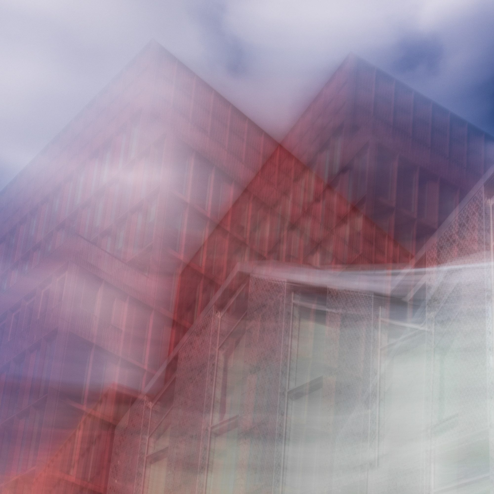 Two red-brick building peaks merging into a pale façade under a stormy sky, from Strata